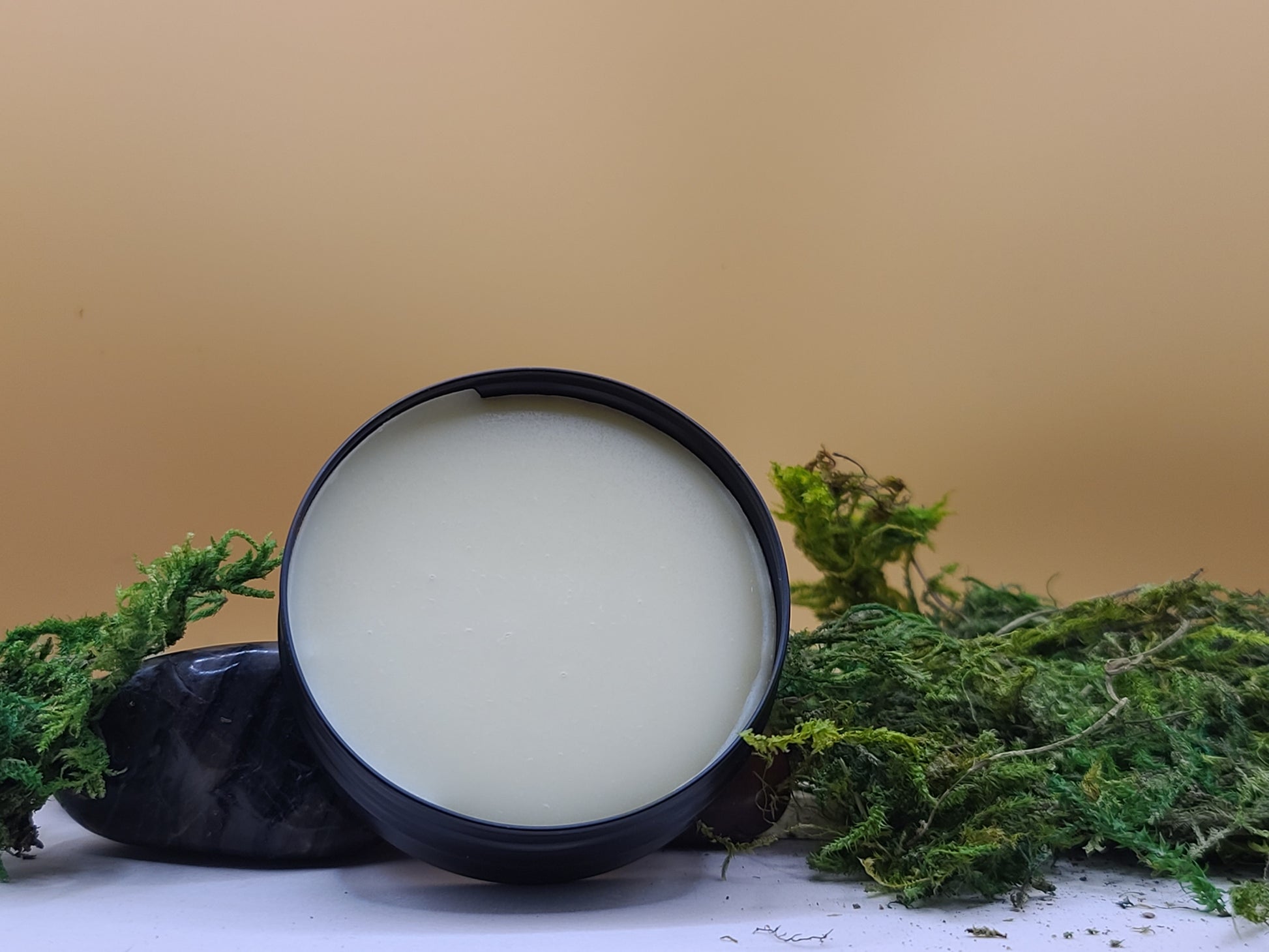 Black container with a white substance surrounded by green foliage on a beige background