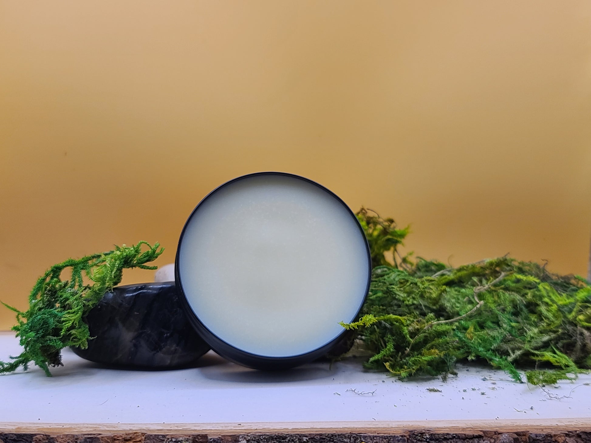 Beard butter with a white center on a wooden surface with green moss, against a beige background.